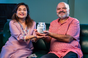 Indian senior couple promoting saving money by putting note in glass jar sitting at home