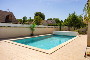 Piscine privée avec margelles en pierre beige, entourée de verdure et équipée d’un rouleau pour bâche, dans un jardin paysager.