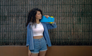 Stylish young woman holding a blue penny board and posing next to a fence