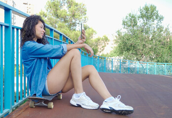 Stylish young woman using a smartphone while sitting on her skateboard on a bridge in an urban environment