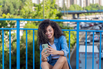 Happy young woman using smartphone while sitting on skateboard on urban bridge with traffic in background