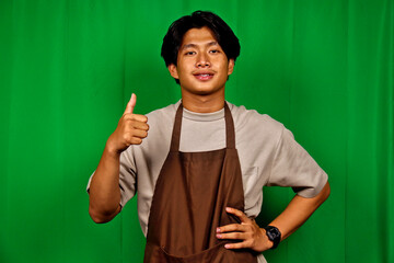 Young Man Wearing Brown Apron Standing in Front of Green Screen Showing Thumbs Up