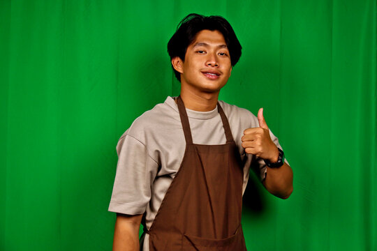 Young Man Wearing Beige T-shirt Brown Apron Giving Thumbs Up in Front of Green Screen - Powered by Adobe