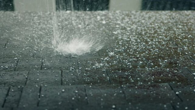 Heavy hail and rain impact a dark asphalt shingle roof, creating splashes and emphasizing the roof's durability and protection against harsh weather conditions.