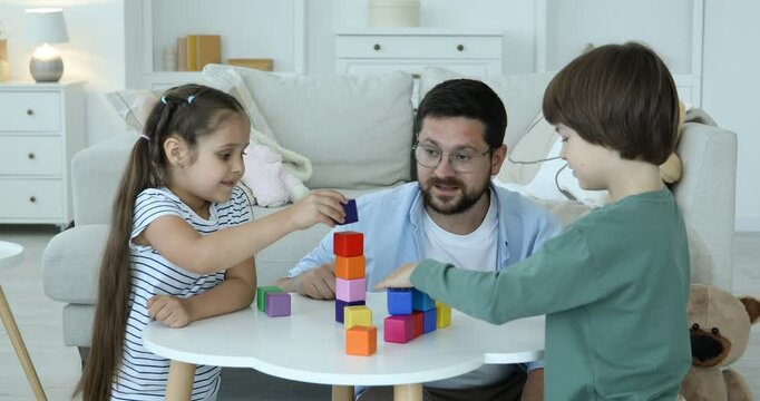 Children and their father building tower with colorful cubes at white table indoors