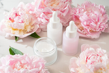 Serum dropper bottle and cream jar with pink peonies, creating a fresh floral skincare flat lay for...