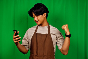 Young Man Wearing Apron Using Smartphone Celebrating Victory in Front of Green Screen