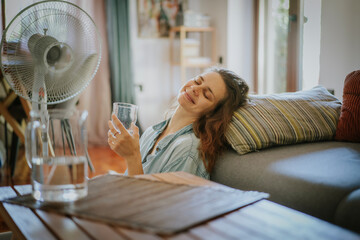 Young woman sitting at home on the floor in front of ventilator and drinking water saving from heat