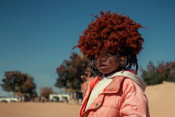A young African girl with dark skin, wearing a vibrant orange wig and pink jacket. A real moment of playful expression, captured in natural light. Perfect for diversity, childhood, and lifestyle theme