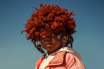 A young African girl with dark skin, wearing a vibrant orange wig and pink jacket. A real moment of playful expression, captured in natural light. Perfect for diversity, childhood, and lifestyle theme