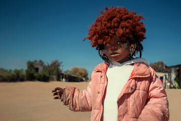 A young African girl with dark skin, wearing a vibrant orange wig and pink jacket. A real moment of playful expression, captured in natural light. Perfect for diversity, childhood, and lifestyle theme