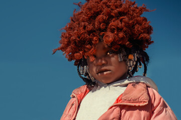 A young African girl with dark skin, wearing a vibrant orange wig and pink jacket. A real moment of playful expression, captured in natural light. Perfect for diversity, childhood, and lifestyle theme
