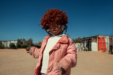 A young African girl with dark skin, wearing a vibrant orange wig and pink jacket. A real moment of playful expression, captured in natural light. Perfect for diversity, childhood, and lifestyle theme