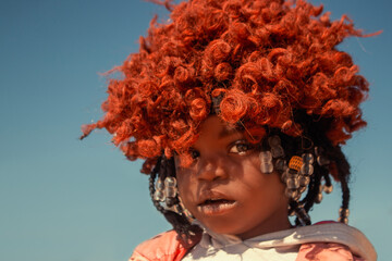 A young African girl with dark skin, wearing a vibrant orange wig and pink jacket. A real moment of playful expression, captured in natural light. Perfect for diversity, childhood, and lifestyle theme