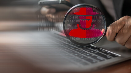 Businessman holding magnifying glass showing hacker icon on laptop keyboard, representing...