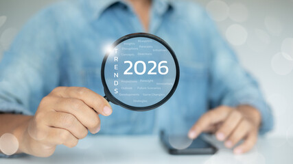 Hand holding magnifying glass focusing on 2026 trends keywords, representing business innovation,...