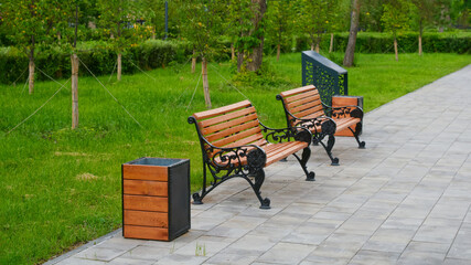 Two empty wooden benches with ornate metal frames and matching trash bins line a paved pathway in a...