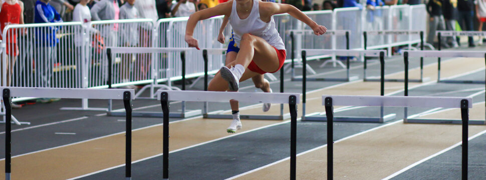 Female Athlete Competes in Indoor Hurdles Event With Spectators in Background