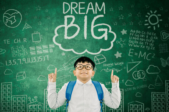 Cheerful Schoolboy with Backpack and Glasses Pointing Up in Front of Inspirational "Dream Big" Chalkboard