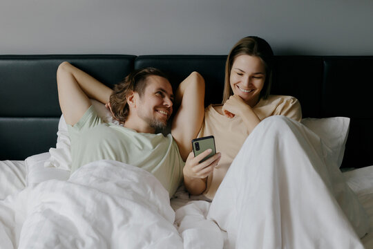 Smiling couple relaxing in bed with smartphone, young caucasian adults enjoying leisure time