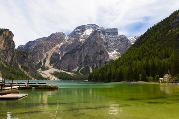 Scenic view of Lake Braies with wooden boats, surrounded by the Seekofel mountain and lush forests in the Dolomites, Italy under a cloudy sky.