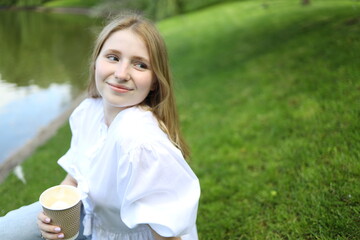 A girl sits on the grass beside a calm lake, smiling softly while holding a warm drink. The sun...