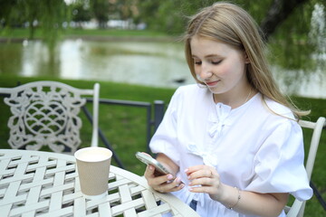 A young woman in a white dress sits at a table with a coffee cup, focused on her smartphone while...