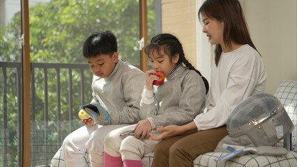 A girl and boy in fencing gear with their mother preparing food and sports equipment