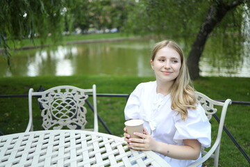 A young woman in a white outfit sits at a table near a pond, holding a warm drink. She enjoys the...