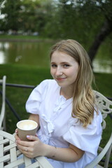 A young woman with long hair sits at a table in a park, smiling while holding a cup of a warm...