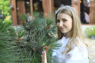 A girl with long blonde hair gently touches the branches of a pine tree, smiling joyfully in a...