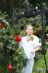 A young woman stands amidst vibrant red roses, smiling while holding a flower. The garden is filled...