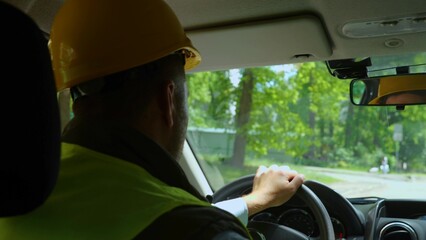 Rear of man worker in yellow helmet and uniform driving a car at a construction site. Close up of...