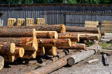 Freshly cut logs are stacked in front of a rustic wooden building with Stacks of pallets in the...