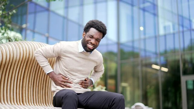 African american businessman feels stomach pain sitting on bench on street near business office building. Sick man has heartburn, gastritis, bloating or poisoning, suffers from spasms and constipation