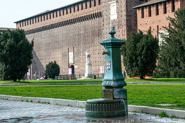 Antique green drinking fountain with the emblem of Milan in front of the historic Sforza Castle. The fountain stands on a cobblestone path surrounded by a well-kept park and lush green lawns.