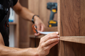 Close-up of man measuring wooden wardrobe with tape measure during furniture assembly, with tools...