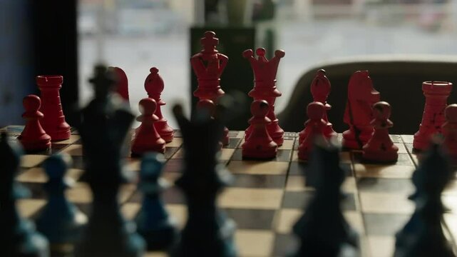 Close-up sequence of a strategic chess match between red and blue pieces on a sunlit chessboard, capturing tension and movement in a thoughtful competitive setting.