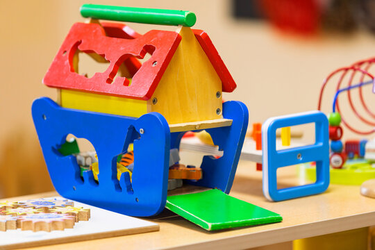 A vibrant wooden toy house, featuring animal cutouts and a shape sorter, sits on a light wooden surface in a kindergarten.