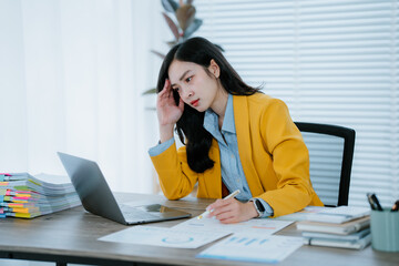 Exhausted young asian businesswoman feeling tired and having a headache while working on a laptop...