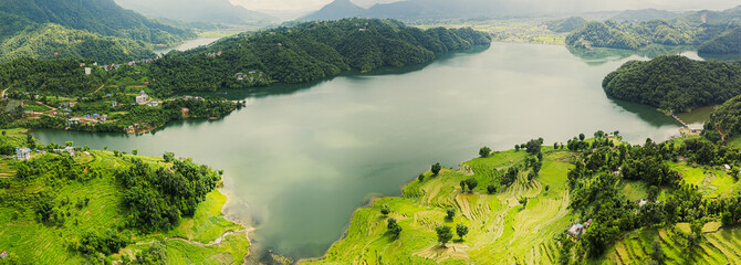 Aerial view of a wide, calm lake surrounded by lush green hills and terraced farmlands, with...