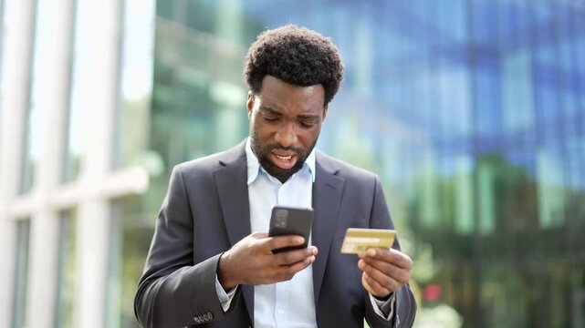 Frustrated african american businessman checks his credit card on phone standing on street near office building. Shocked worker in formal suit faced fraud, money was stolen from his account, deception