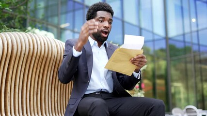 Smiling satisfied african american businessman in suit reading letter with great news on street near business office building. Worker is celebrating success, happy to receive a pleasant notification