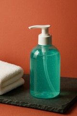 A bottle of teal liquid soap with a pump dispenser sits on a dark gray surface next to a folded white towel against a burnt orange background. 