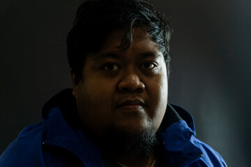 Bearded Indonesian man in his 30s wearing a shiny blue nylon jacket against black background,...