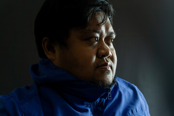 Bearded Indonesian man in his 30s wearing a shiny blue nylon jacket against black background,...