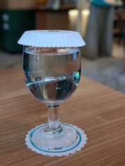 A glass of water with a white paper cover sits on a wooden table, with a decorative coaster underneath