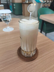 An iced coffee drink with froth and a stirrer sits on a wooden coaster, next to a glass of water, on a wooden table.