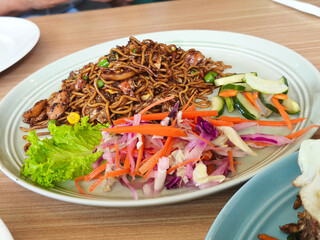 
A plate of Indonesian Mie Goreng or fried noodles with chicken, vegetables, pickled cucumber and carrot, and fresh salad, on a wooden table in Surabaya.