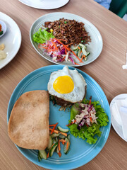 A blue plate holds Indonesian fried rice with a sunny-side-up egg, a large prawn cracker, pickled vegetables, and fresh salad on a wooden table.